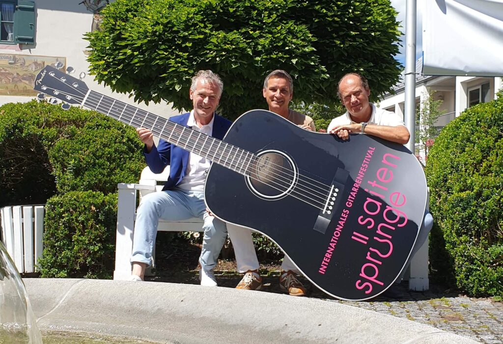 Wiedemann, Jahn und Erkes auf Bank vor Brunnen mit großer Saitensprünge-Dekogitarre