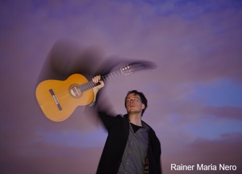 Rainer-Maria-Nero-gallerie Rainer Maria Nero hält Gitarre hoch vor lila Hintergrund