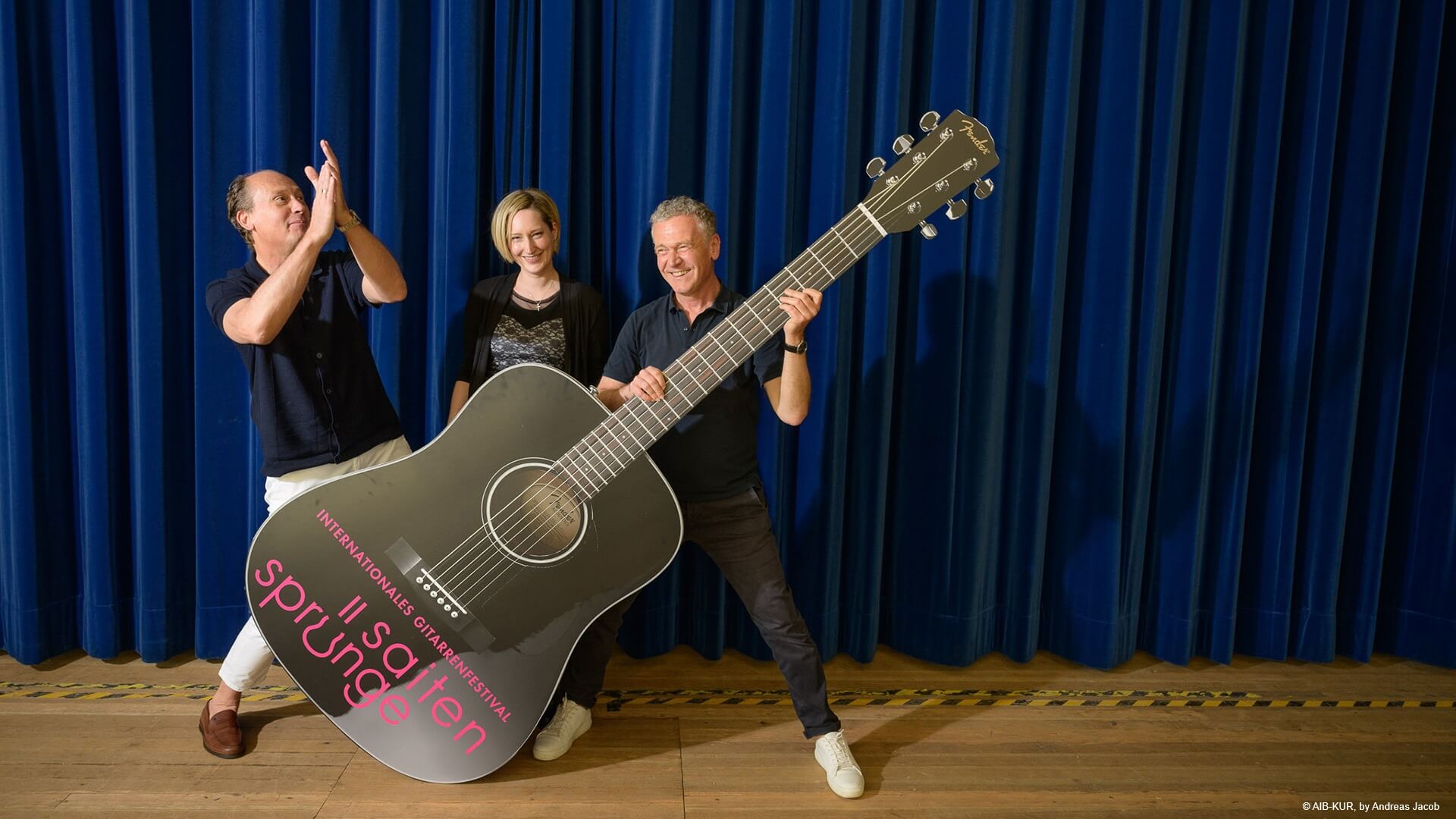 Das Festival-Team: Erkes, Stuber und Wiedemann mit großer Gitarre auf Bühne